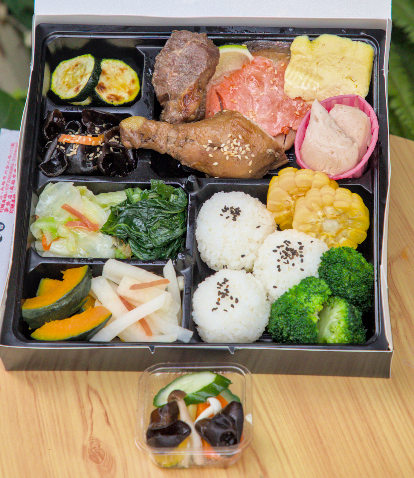 紅蕃茄客製化便當,無炸物無麩質的健康選擇 紅蕃茄客製化便當的俯視圖,便當盒分格擺放著多樣主菜與配菜,如滷雞腿、生食級鮭魚、烤櫛瓜、黑木耳等,色彩繽紛,看起來既美味又健康。